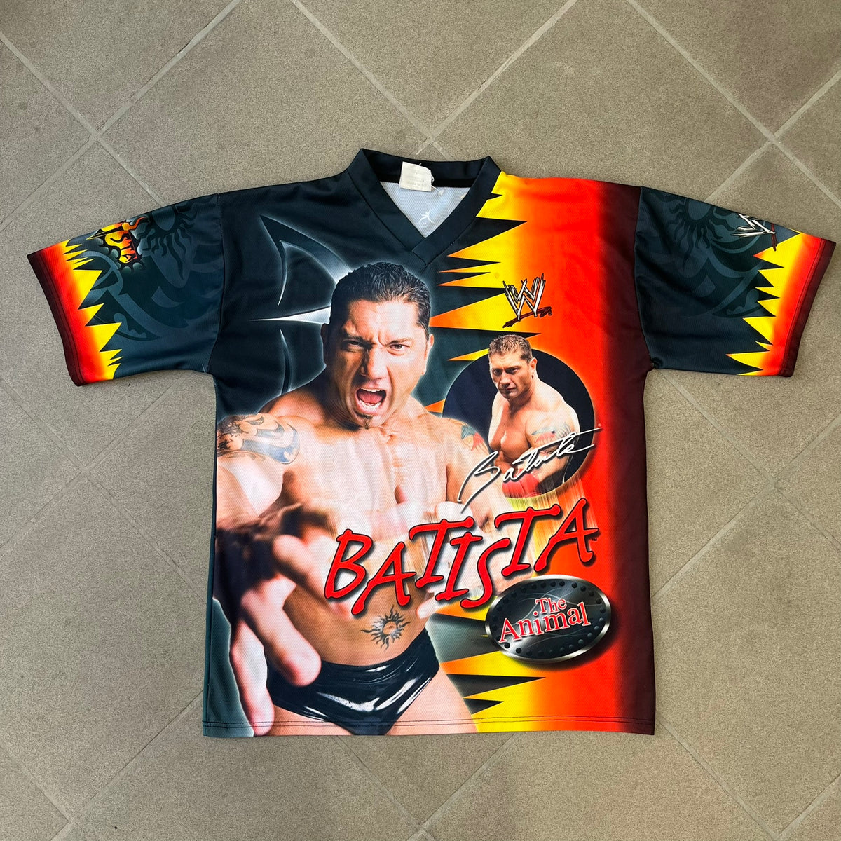 WWE Batista 2007 Kalciomania AOP Tee features an all-over print design with red, black, and white graphics throughout. The front includes Two Batista graphics with red&nbsp; “Batista” text, WWE logos, and additional printed details. The back features Batista with Unleashed Batista text graphic. The sleeves include a WWE logo and branding. Finished with a Kalciomania licensed tag.