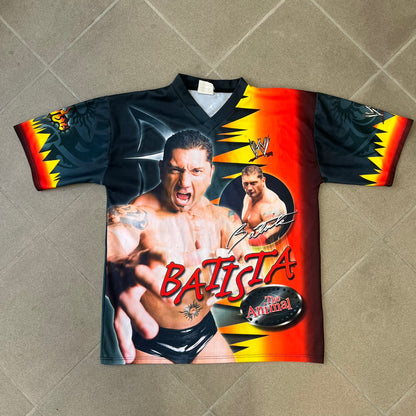 WWE Batista 2007 Kalciomania AOP Tee features an all-over print design with red, black, and white graphics throughout. The front includes Two Batista graphics with red&nbsp; “Batista” text, WWE logos, and additional printed details. The back features Batista with Unleashed Batista text graphic. The sleeves include a WWE logo and branding. Finished with a Kalciomania licensed tag.