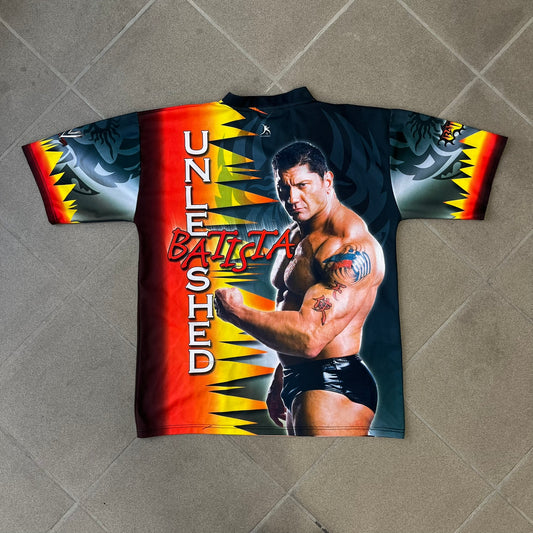 WWE Batista 2007 Kalciomania AOP Tee features an all-over print design with red, black, and white graphics throughout. The front includes Two Batista graphics with red&nbsp; “Batista” text, WWE logos, and additional printed details. The back features Batista with Unleashed Batista text graphic. The sleeves include a WWE logo and branding. Finished with a Kalciomania licensed tag.