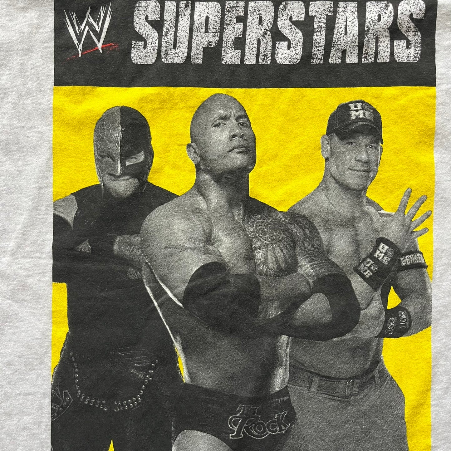Vintage WWE Superstars Tee in white, highlighted by a large front print of wrestling icons The Rock, Rey Mysterio, and John Cena over a yellow base. Features the WWE logo and “Superstars” branding, printed on a Fruit of the Loom blank.
