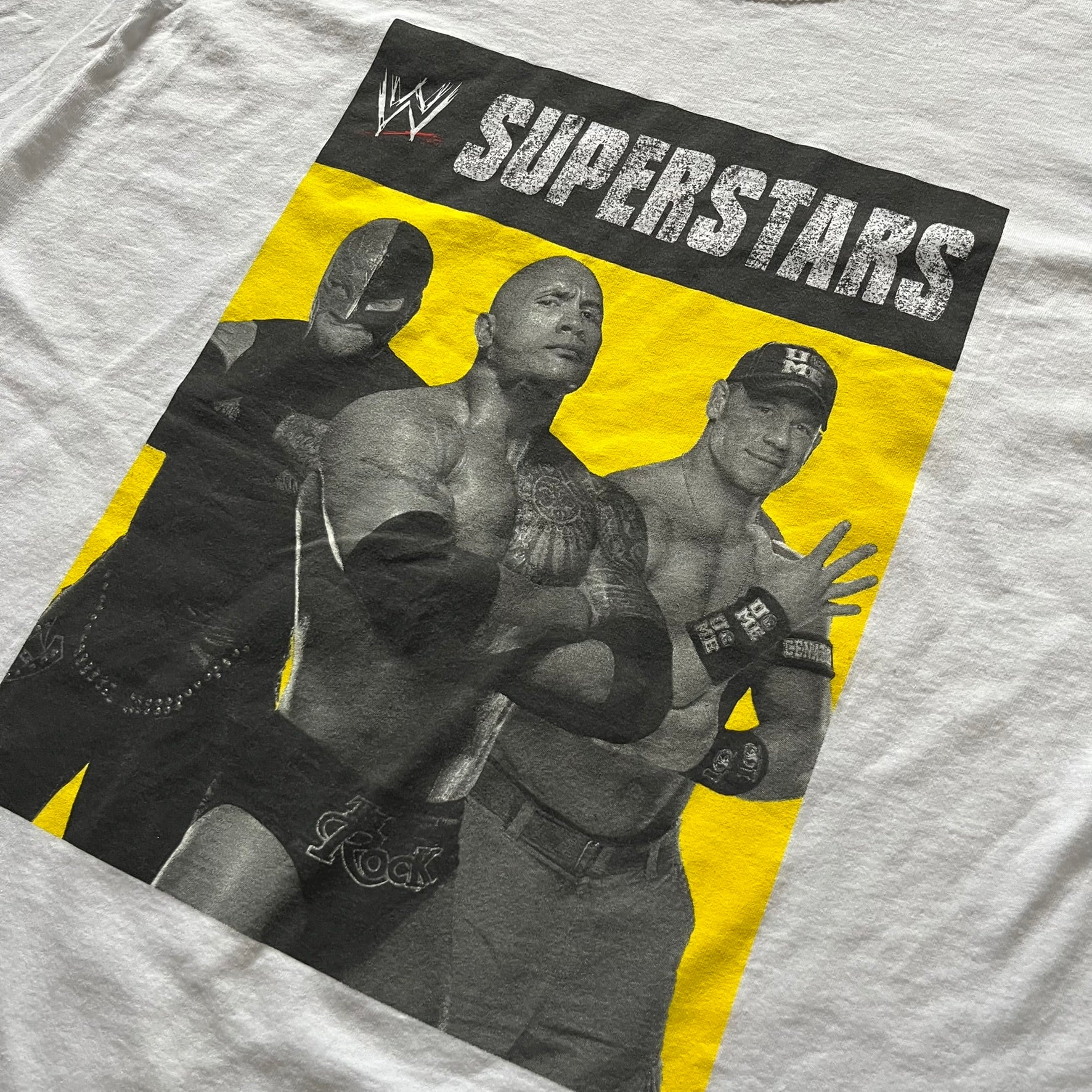Vintage WWE Superstars Tee in white, highlighted by a large front print of wrestling icons The Rock, Rey Mysterio, and John Cena over a yellow base. Features the WWE logo and “Superstars” branding, printed on a Fruit of the Loom blank.