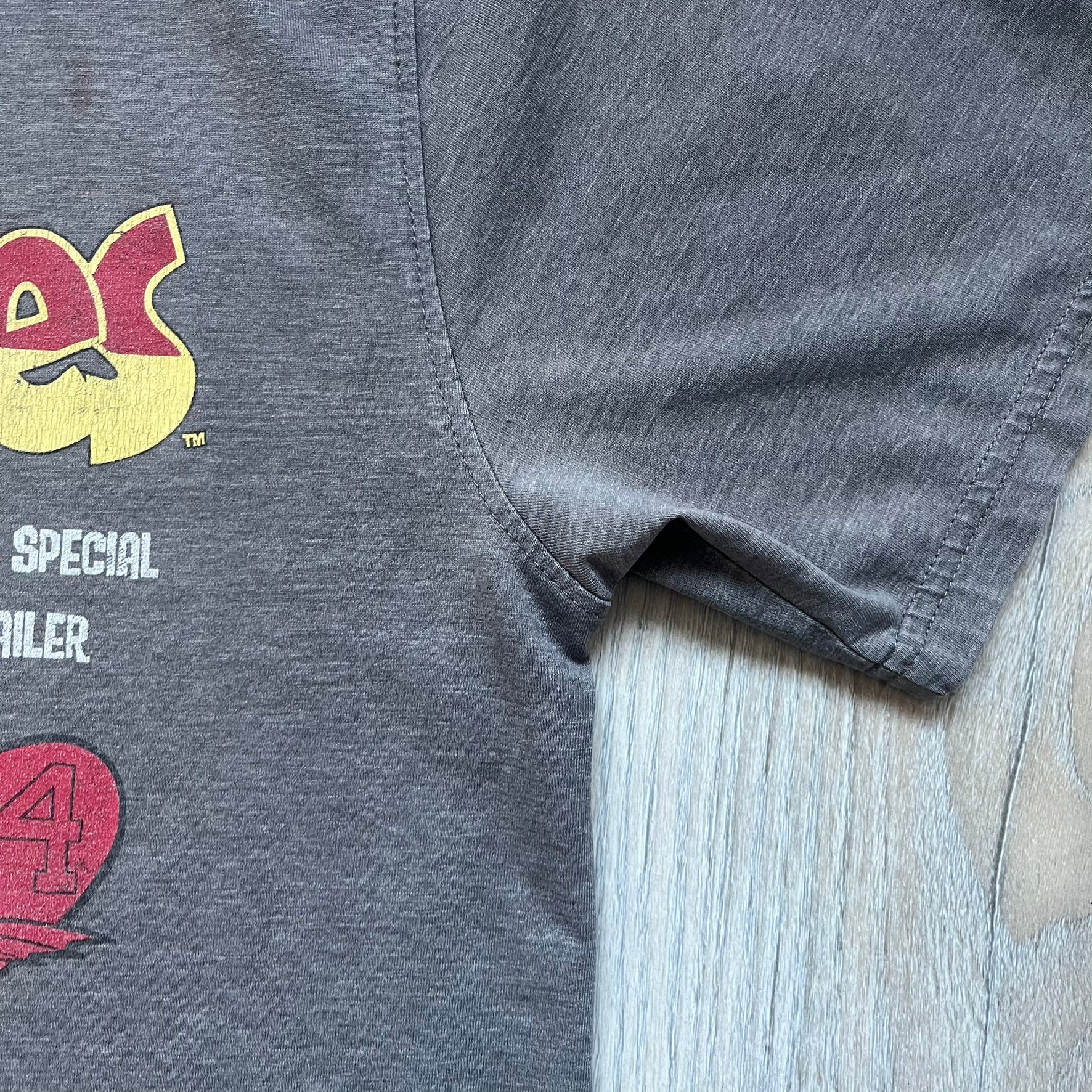 Wacky Races Gray 2010 Tee is a short-sleeve t-shirt featuring a front graphic with red and yellow “Wacky Races” text at the top. The design shows three cars at the starting line: the Army Surplus Special, the Crimson Haybaler, and the Mean Machine, each labeled with small white text next to the vehicles. The tee includes a purple Wacky Races neck tag with Cartoon Network 2010 licensing.