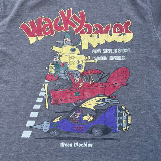 Wacky Races Gray 2010 Tee is a short-sleeve t-shirt featuring a front graphic with red and yellow “Wacky Races” text at the top. The design shows three cars at the starting line: the Army Surplus Special, the Crimson Haybaler, and the Mean Machine, each labeled with small white text next to the vehicles. The tee includes a purple Wacky Races neck tag with Cartoon Network 2010 licensing.