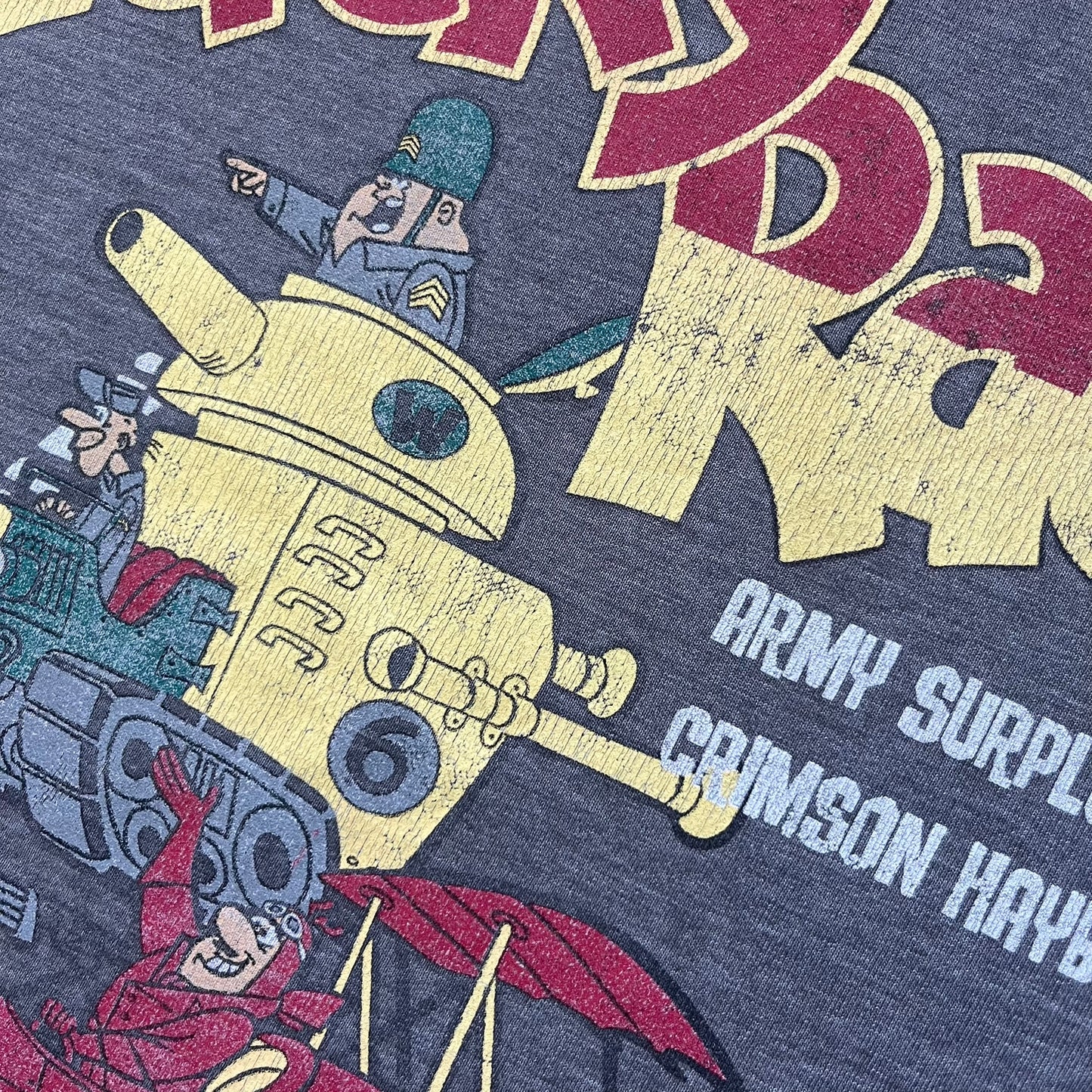 Wacky Races Gray 2010 Tee is a short-sleeve t-shirt featuring a front graphic with red and yellow “Wacky Races” text at the top. The design shows three cars at the starting line: the Army Surplus Special, the Crimson Haybaler, and the Mean Machine, each labeled with small white text next to the vehicles. The tee includes a purple Wacky Races neck tag with Cartoon Network 2010 licensing.