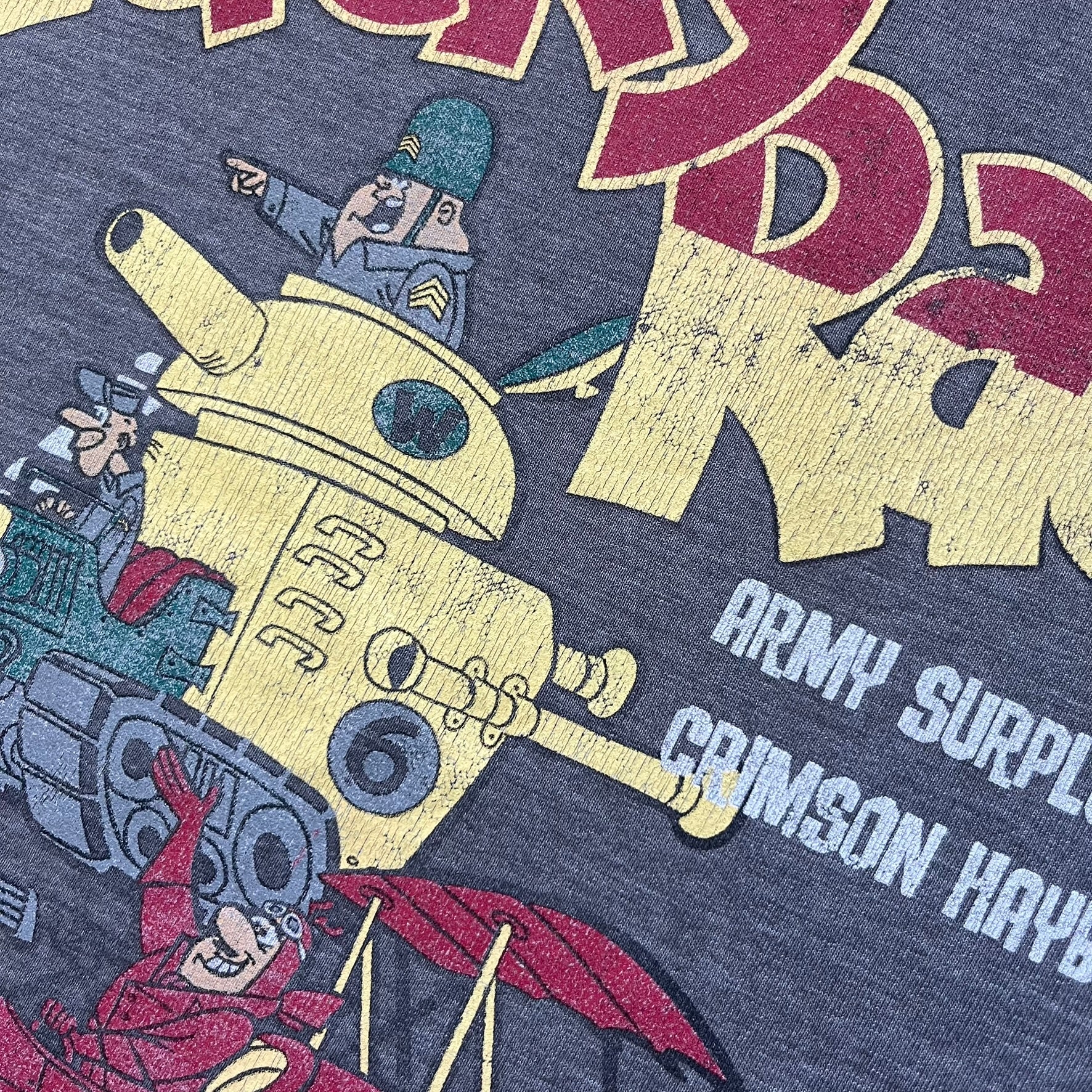 Wacky Races Gray 2010 Tee is a short-sleeve t-shirt featuring a front graphic with red and yellow “Wacky Races” text at the top. The design shows three cars at the starting line: the Army Surplus Special, the Crimson Haybaler, and the Mean Machine, each labeled with small white text next to the vehicles. The tee includes a purple Wacky Races neck tag with Cartoon Network 2010 licensing.