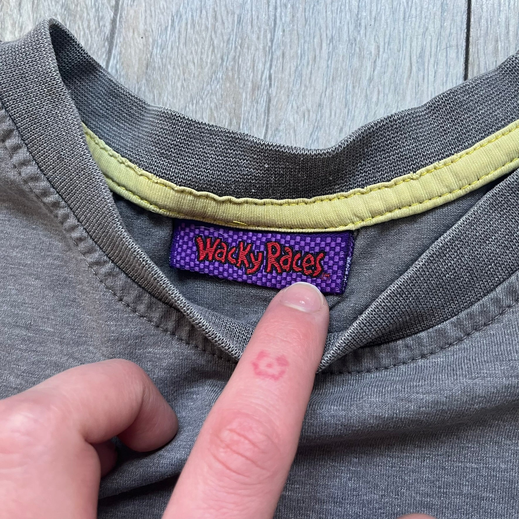 Wacky Races Gray 2010 Tee is a short-sleeve t-shirt featuring a front graphic with red and yellow “Wacky Races” text at the top. The design shows three cars at the starting line: the Army Surplus Special, the Crimson Haybaler, and the Mean Machine, each labeled with small white text next to the vehicles. The tee includes a purple Wacky Races neck tag with Cartoon Network 2010 licensing.