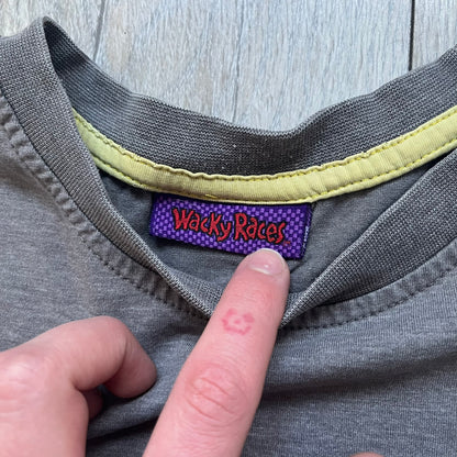 Wacky Races Gray 2010 Tee is a short-sleeve t-shirt featuring a front graphic with red and yellow “Wacky Races” text at the top. The design shows three cars at the starting line: the Army Surplus Special, the Crimson Haybaler, and the Mean Machine, each labeled with small white text next to the vehicles. The tee includes a purple Wacky Races neck tag with Cartoon Network 2010 licensing.