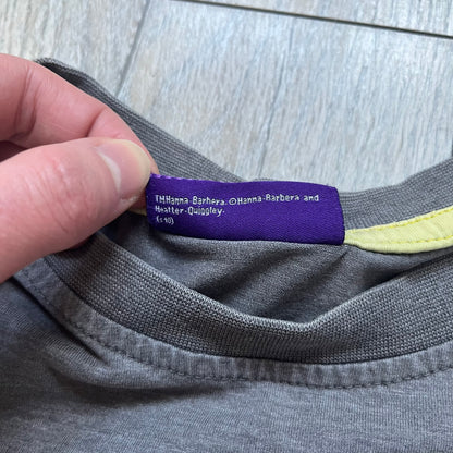 Wacky Races Gray 2010 Tee is a short-sleeve t-shirt featuring a front graphic with red and yellow “Wacky Races” text at the top. The design shows three cars at the starting line: the Army Surplus Special, the Crimson Haybaler, and the Mean Machine, each labeled with small white text next to the vehicles. The tee includes a purple Wacky Races neck tag with Cartoon Network 2010 licensing.