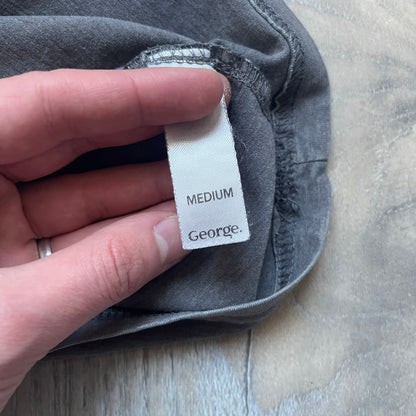 Wacky Races Gray 2010 Tee is a short-sleeve t-shirt featuring a front graphic with red and yellow “Wacky Races” text at the top. The design shows three cars at the starting line: the Army Surplus Special, the Crimson Haybaler, and the Mean Machine, each labeled with small white text next to the vehicles. The tee includes a purple Wacky Races neck tag with Cartoon Network 2010 licensing.