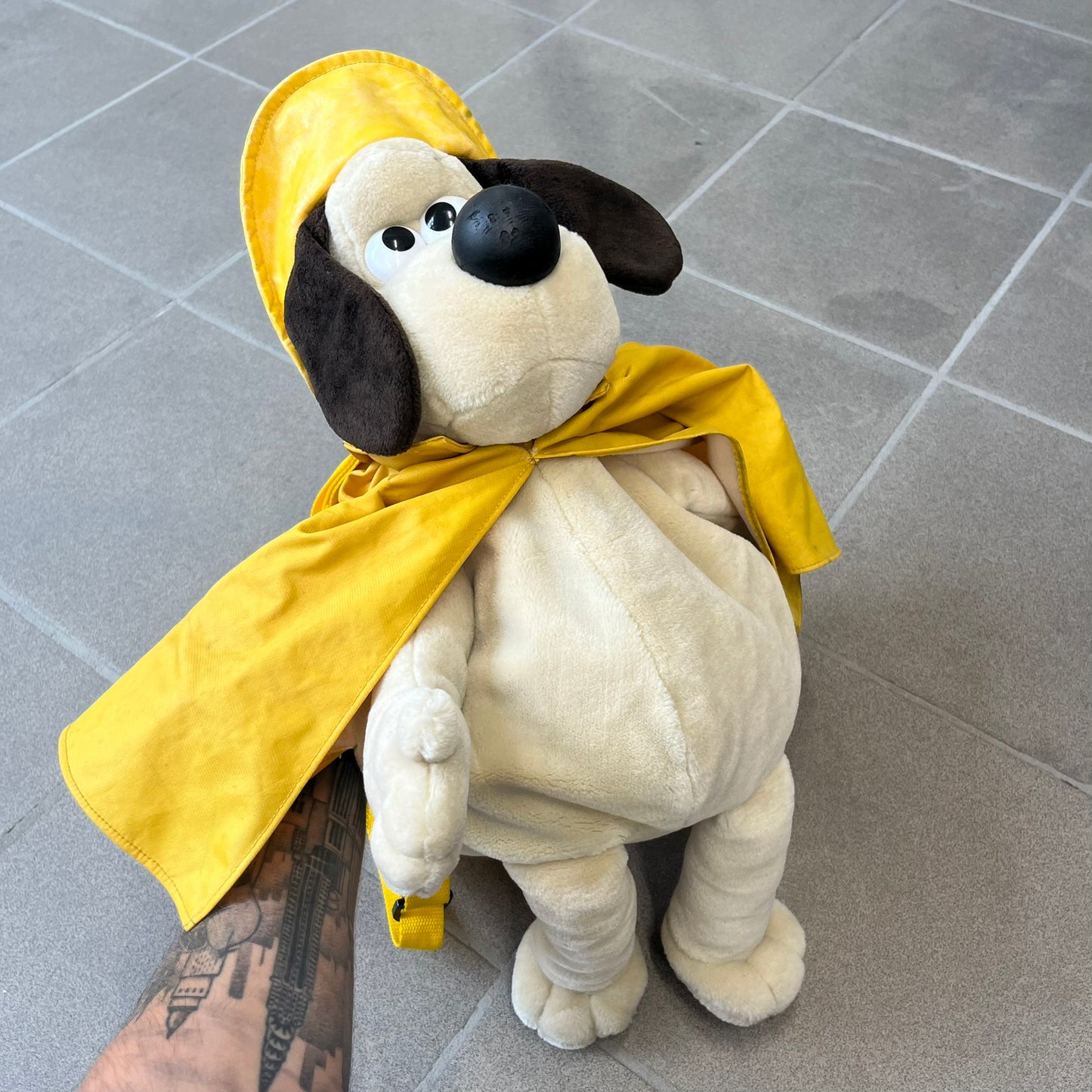 The Wallace and Gromit Gromit Plush Backpack is a 1990s licensed item produced by Aardman Animations. It features Gromit dressed in his bright yellow PVC raincoat and matching hat, as seen in The Wrong Trousers. The backpack is made from soft plush fabric with plastic eyes, a hard black nose, and a genuine stamp confirming official production. The bag opens from the side and includes adjustable shoulder straps for wearability.