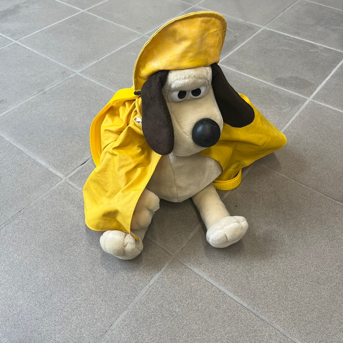 The Wallace and Gromit Gromit Plush Backpack is a 1990s licensed item produced by Aardman Animations. It features Gromit dressed in his bright yellow PVC raincoat and matching hat, as seen in The Wrong Trousers. The backpack is made from soft plush fabric with plastic eyes, a hard black nose, and a genuine stamp confirming official production. The bag opens from the side and includes adjustable shoulder straps for wearability.