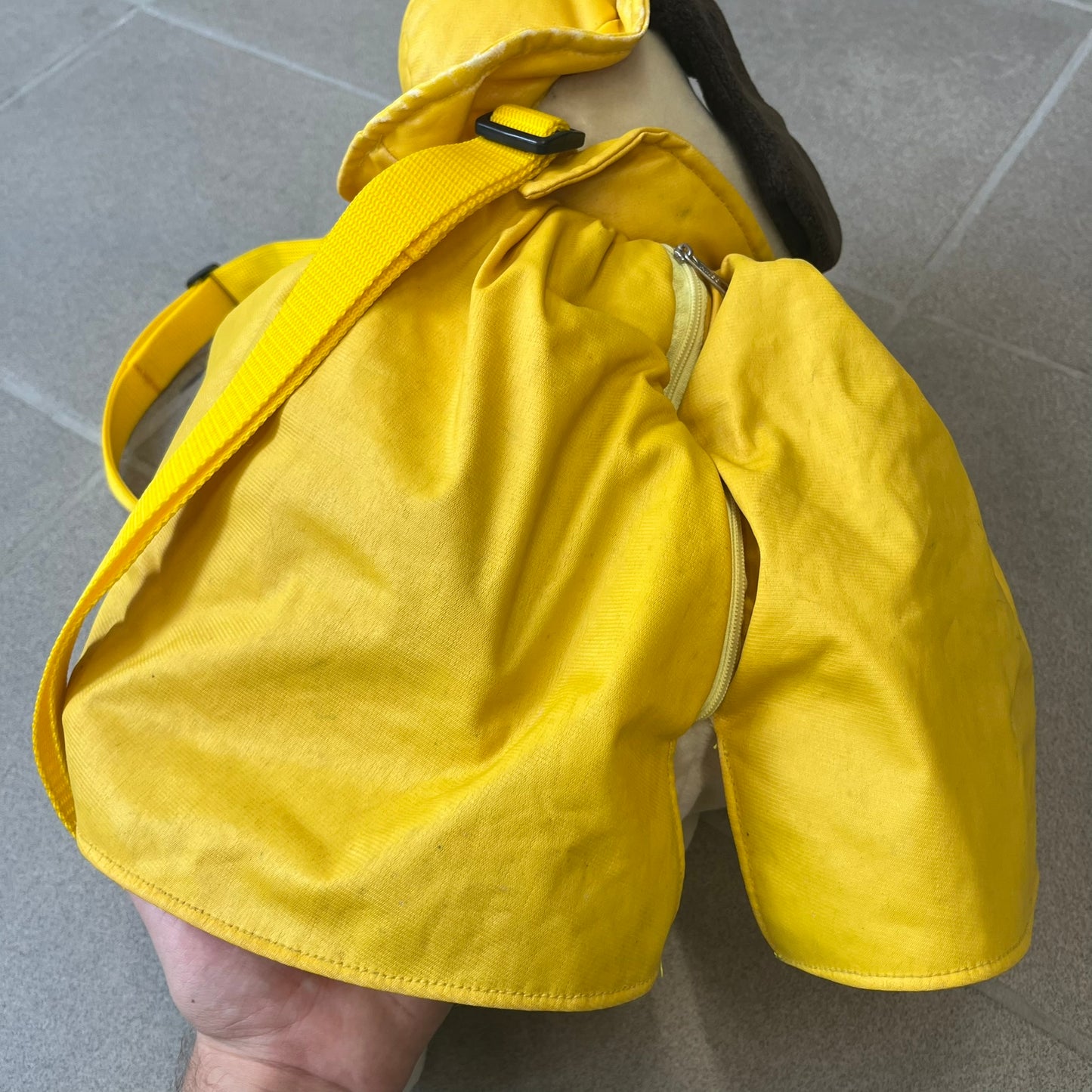 The Wallace and Gromit Gromit Plush Backpack is a 1990s licensed item produced by Aardman Animations. It features Gromit dressed in his bright yellow PVC raincoat and matching hat, as seen in The Wrong Trousers. The backpack is made from soft plush fabric with plastic eyes, a hard black nose, and a genuine stamp confirming official production. The bag opens from the side and includes adjustable shoulder straps for wearability.