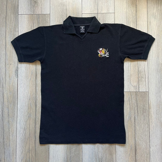 Warner Bros 1999 Waffle Knit Black Polo is a black polo shirt featuring a textured waffle knit construction. The left chest includes an embroidered graphic of Taz peeking behind a Warner Bros logo, with Bugs Bunny and Tweety depicted running away. The shirt has an official licensed neck tag and dates to 1999.
