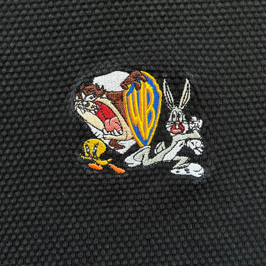 Warner Bros 1999 Waffle Knit Black Polo is a black polo shirt featuring a textured waffle knit construction. The left chest includes an embroidered graphic of Taz peeking behind a Warner Bros logo, with Bugs Bunny and Tweety depicted running away. The shirt has an official licensed neck tag and dates to 1999.