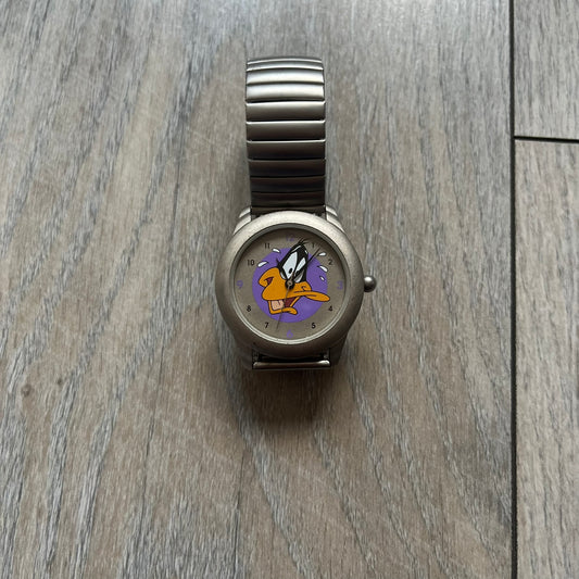 Warner Bros Daffy Duck 1997 Analog Watch is a stainless steel watch with a matte finish and a stretch expansion bracelet made from connected metal links. The dial features black and purple numerals with a graphic of Daffy Duck shown sweating in front of a purple circle. The caseback includes Warner Bros 1997 licensing.