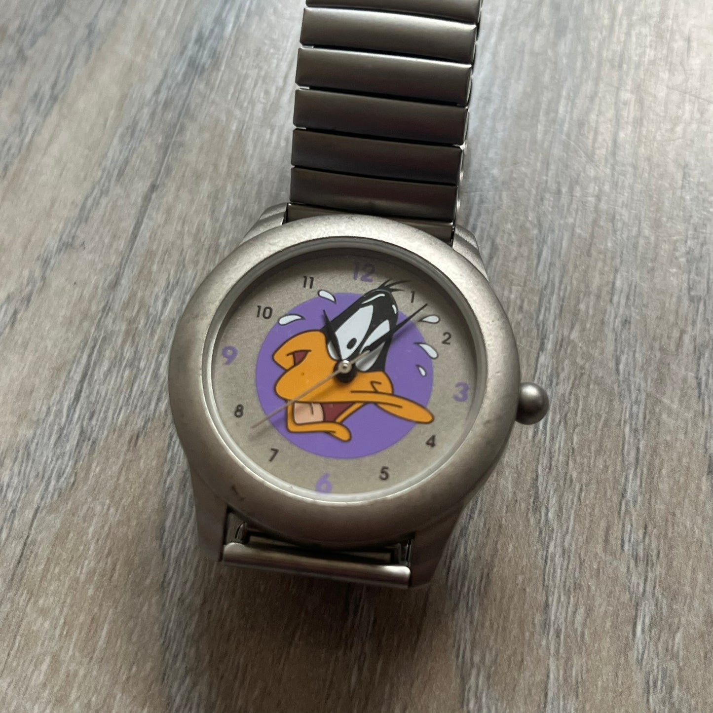Warner Bros Daffy Duck 1997 Analog Watch is a stainless steel watch with a matte finish and a stretch expansion bracelet made from connected metal links. The dial features black and purple numerals with a graphic of Daffy Duck shown sweating in front of a purple circle. The caseback includes Warner Bros 1997 licensing.