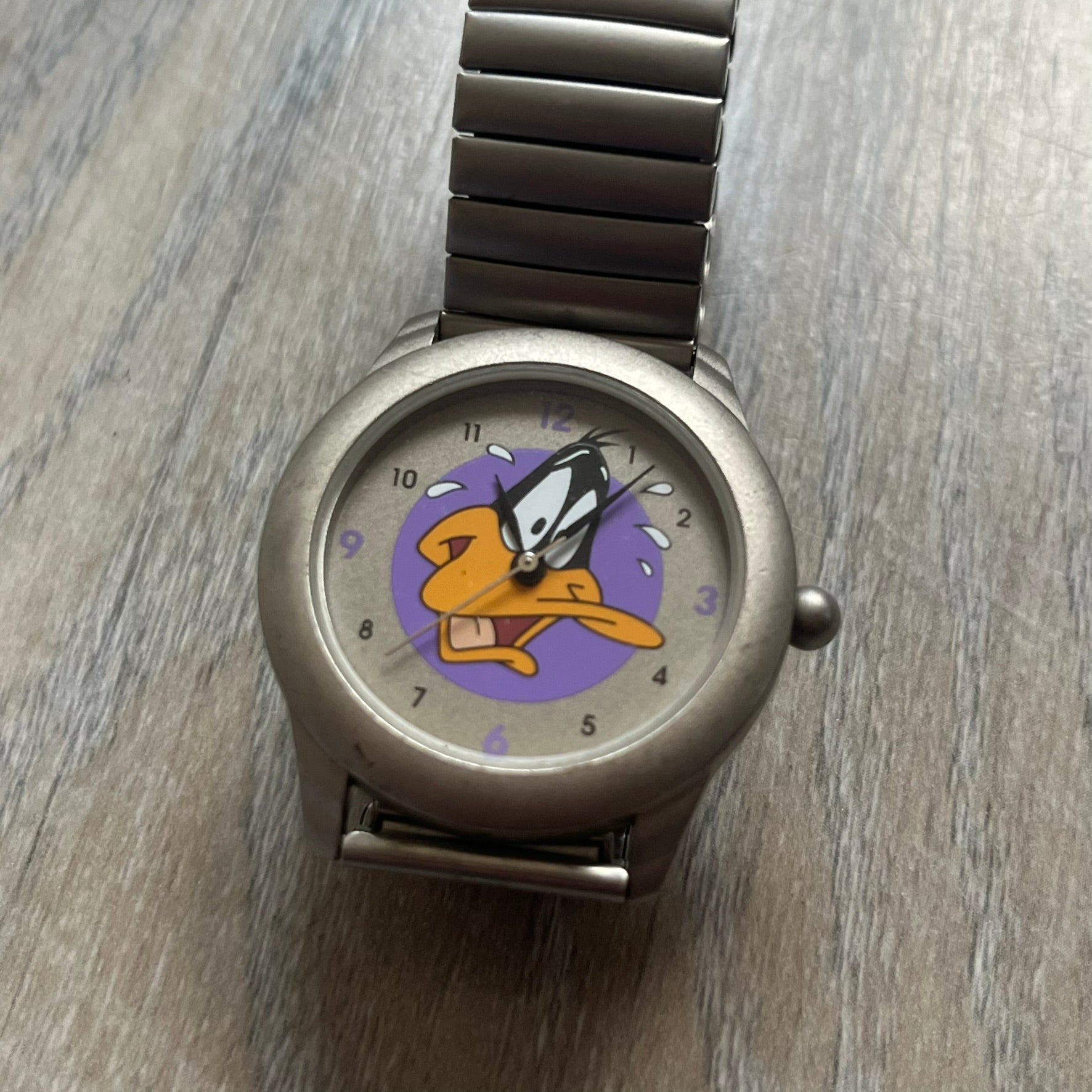 Warner Bros Daffy Duck 1997 Analog Watch is a stainless steel watch with a matte finish and a stretch expansion bracelet made from connected metal links. The dial features black and purple numerals with a graphic of Daffy Duck shown sweating in front of a purple circle. The caseback includes Warner Bros 1997 licensing.