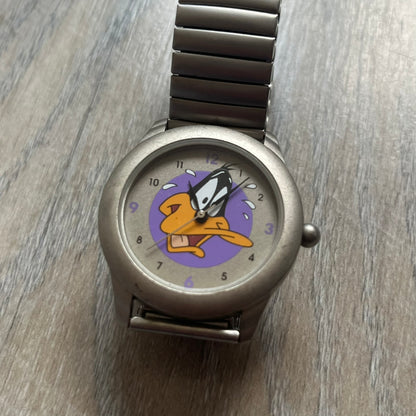 Warner Bros Daffy Duck 1997 Analog Watch is a stainless steel watch with a matte finish and a stretch expansion bracelet made from connected metal links. The dial features black and purple numerals with a graphic of Daffy Duck shown sweating in front of a purple circle. The caseback includes Warner Bros 1997 licensing.