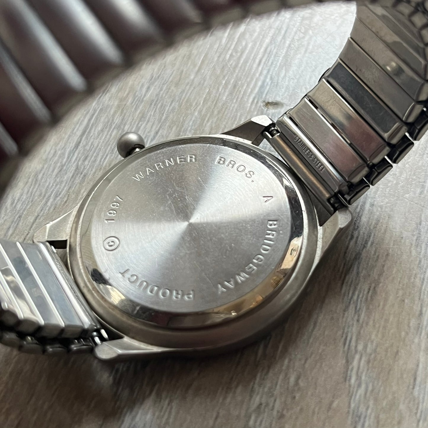Warner Bros Daffy Duck 1997 Analog Watch is a stainless steel watch with a matte finish and a stretch expansion bracelet made from connected metal links. The dial features black and purple numerals with a graphic of Daffy Duck shown sweating in front of a purple circle. The caseback includes Warner Bros 1997 licensing.