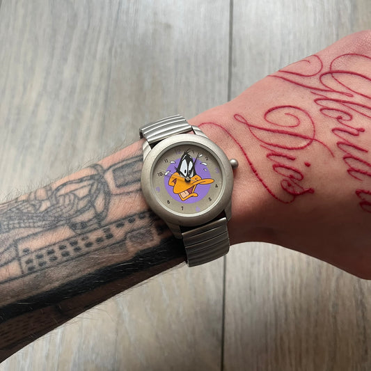 Warner Bros Daffy Duck 1997 Analog Watch is a stainless steel watch with a matte finish and a stretch expansion bracelet made from connected metal links. The dial features black and purple numerals with a graphic of Daffy Duck shown sweating in front of a purple circle. The caseback includes Warner Bros 1997 licensing.