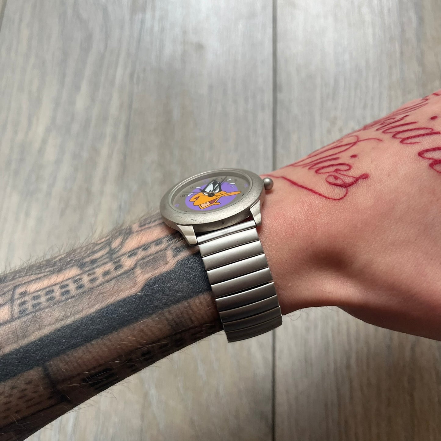Warner Bros Daffy Duck 1997 Analog Watch is a stainless steel watch with a matte finish and a stretch expansion bracelet made from connected metal links. The dial features black and purple numerals with a graphic of Daffy Duck shown sweating in front of a purple circle. The caseback includes Warner Bros 1997 licensing.