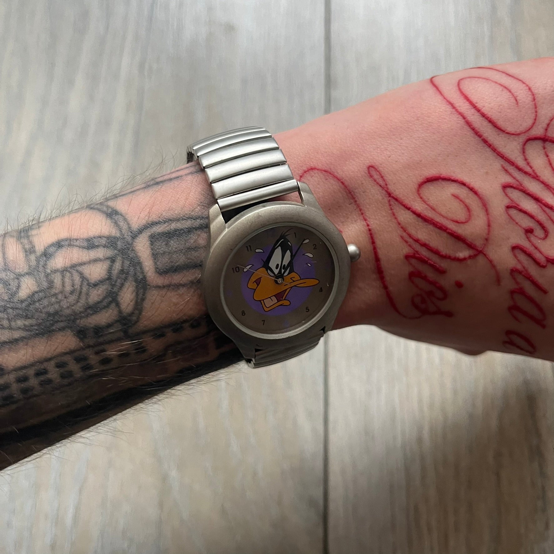 Warner Bros Daffy Duck 1997 Analog Watch is a stainless steel watch with a matte finish and a stretch expansion bracelet made from connected metal links. The dial features black and purple numerals with a graphic of Daffy Duck shown sweating in front of a purple circle. The caseback includes Warner Bros 1997 licensing.
