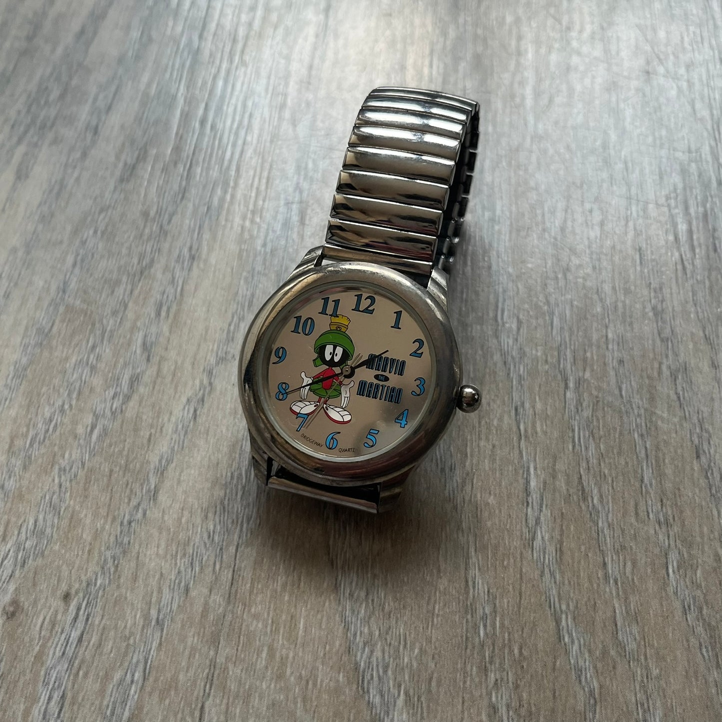 Warner Bros Marvin the Martian 1996 Analog Watch is a stainless steel watch with a shiny finish and a stretch expansion bracelet made from connected metal links. The dial is metallic and reflective with blue numerals and a graphic of Marvin the Martian standing with his name printed beside him. The caseback includes Warner Bros 1996 licensing.