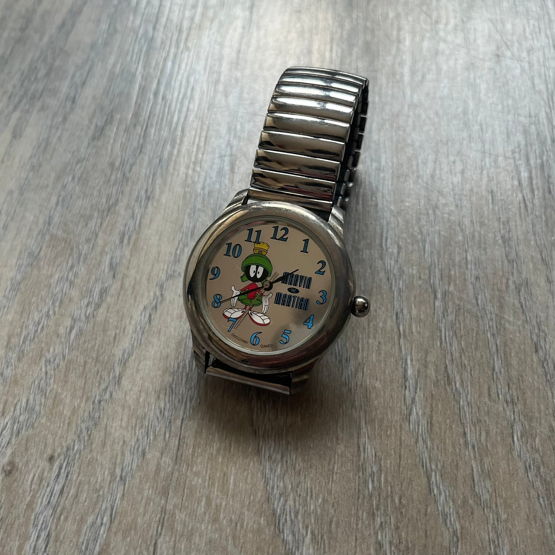 Warner Bros Marvin the Martian 1996 Analog Watch is a stainless steel watch with a shiny finish and a stretch expansion bracelet made from connected metal links. The dial is metallic and reflective with blue numerals and a graphic of Marvin the Martian standing with his name printed beside him. The caseback includes Warner Bros 1996 licensing.