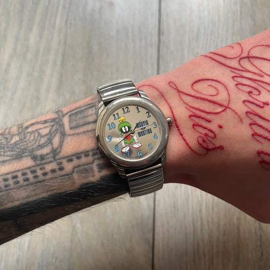 Warner Bros Marvin the Martian 1996 Analog Watch is a stainless steel watch with a shiny finish and a stretch expansion bracelet made from connected metal links. The dial is metallic and reflective with blue numerals and a graphic of Marvin the Martian standing with his name printed beside him. The caseback includes Warner Bros 1996 licensing.