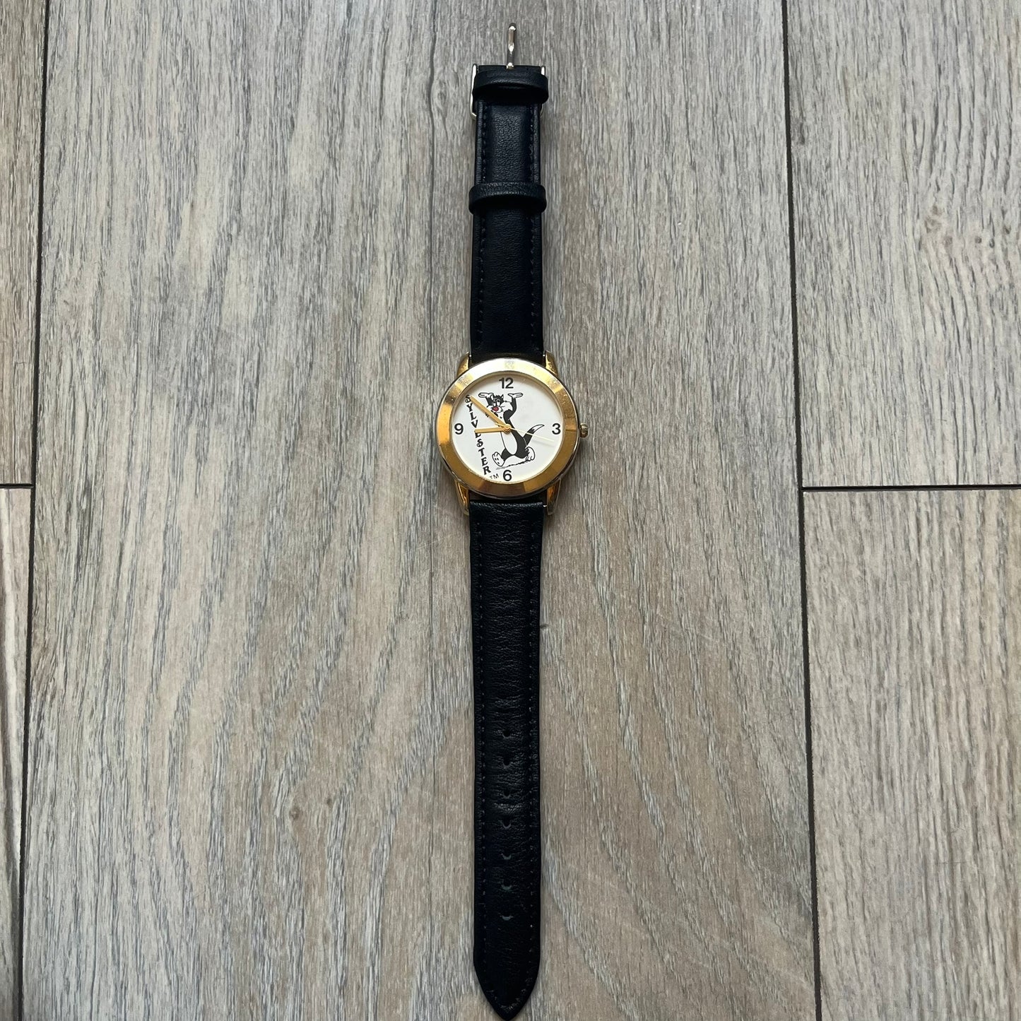 Warner Bros Sylvester 1990 Analog Watch features a gold-tone case with a white dial and black leather strap marked as genuine leather. The dial displays Sylvester with vertical “Sylvester” text, along with hour markers at 12, 3, 6, and 9. The watch uses a Japan movement and includes Warner Bros 1990 licensing engraved on the caseback