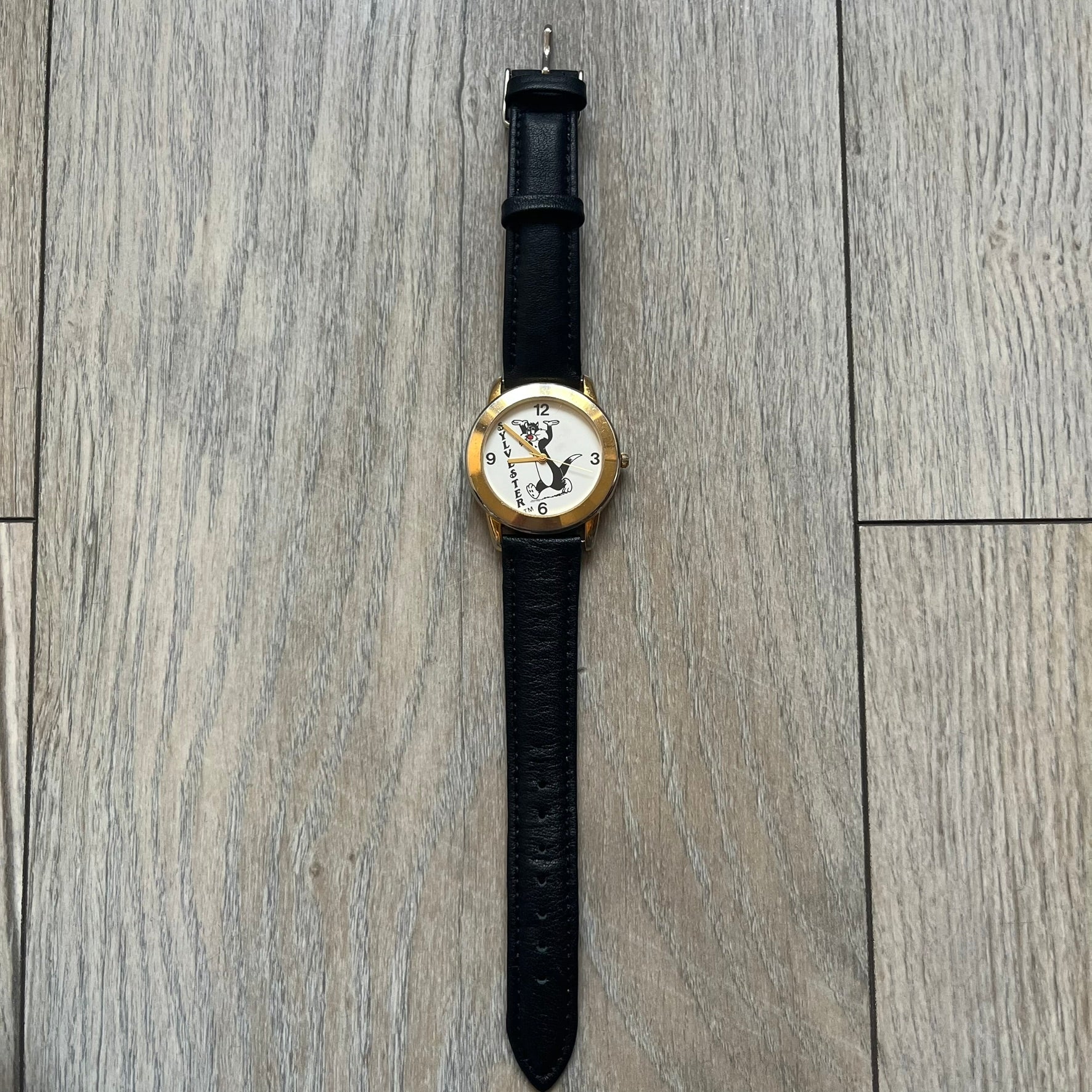 Warner Bros Sylvester 1990 Analog Watch features a gold-tone case with a white dial and black leather strap marked as genuine leather. The dial displays Sylvester with vertical “Sylvester” text, along with hour markers at 12, 3, 6, and 9. The watch uses a Japan movement and includes Warner Bros 1990 licensing engraved on the caseback