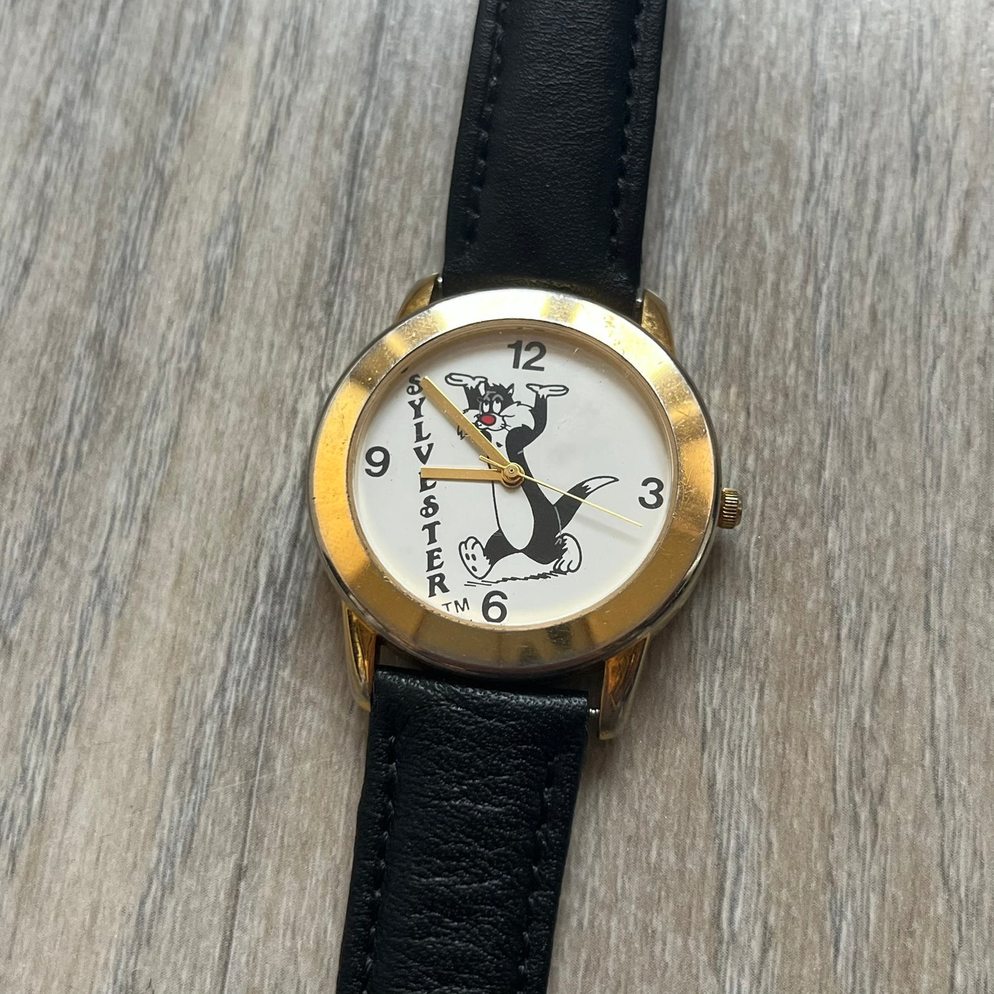 Warner Bros Sylvester 1990 Analog Watch features a gold-tone case with a white dial and black leather strap marked as genuine leather. The dial displays Sylvester with vertical “Sylvester” text, along with hour markers at 12, 3, 6, and 9. The watch uses a Japan movement and includes Warner Bros 1990 licensing engraved on the caseback