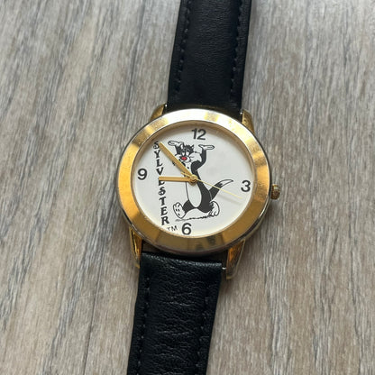 Warner Bros Sylvester 1990 Analog Watch features a gold-tone case with a white dial and black leather strap marked as genuine leather. The dial displays Sylvester with vertical “Sylvester” text, along with hour markers at 12, 3, 6, and 9. The watch uses a Japan movement and includes Warner Bros 1990 licensing engraved on the caseback