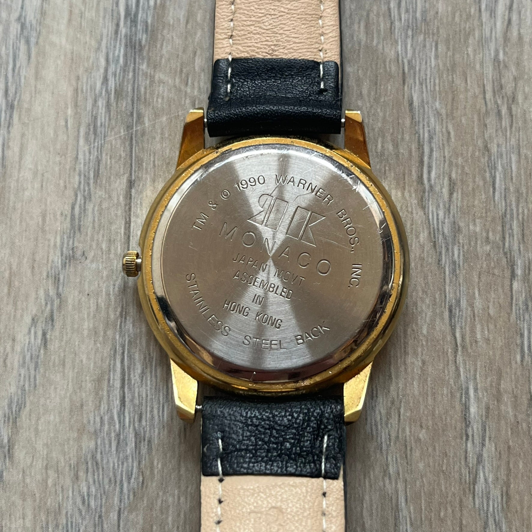 Warner Bros Sylvester 1990 Analog Watch features a gold-tone case with a white dial and black leather strap marked as genuine leather. The dial displays Sylvester with vertical “Sylvester” text, along with hour markers at 12, 3, 6, and 9. The watch uses a Japan movement and includes Warner Bros 1990 licensing engraved on the caseback