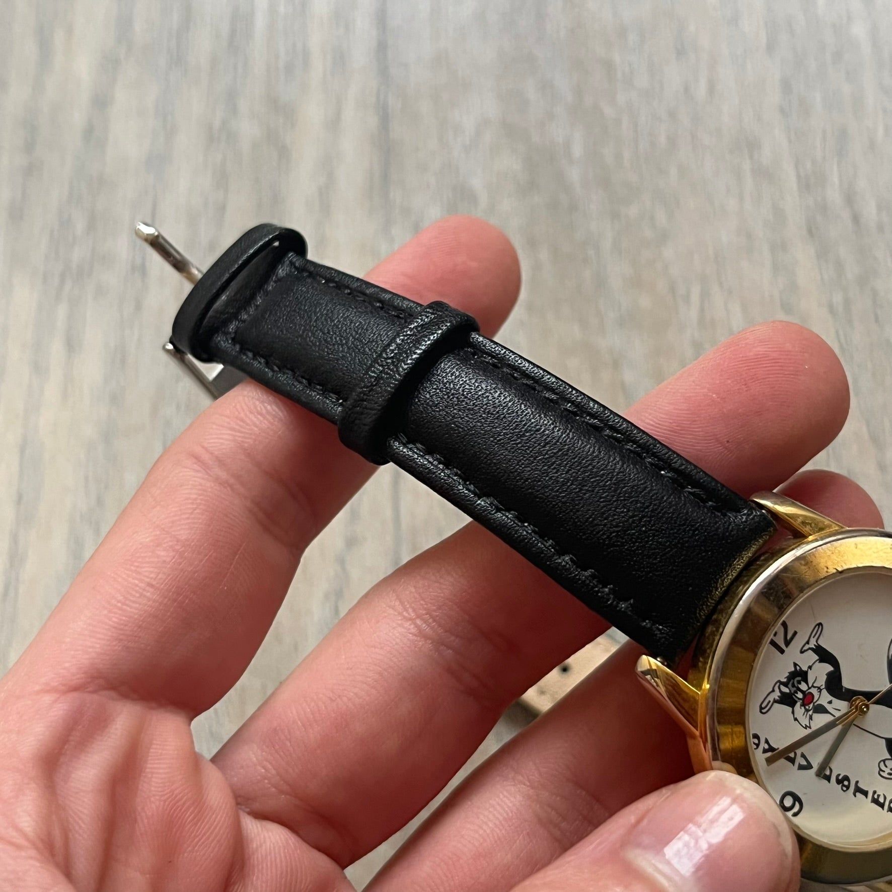 Warner Bros Sylvester 1990 Analog Watch features a gold-tone case with a white dial and black leather strap marked as genuine leather. The dial displays Sylvester with vertical “Sylvester” text, along with hour markers at 12, 3, 6, and 9. The watch uses a Japan movement and includes Warner Bros 1990 licensing engraved on the caseback