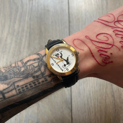 Warner Bros Sylvester 1990 Analog Watch features a gold-tone case with a white dial and black leather strap marked as genuine leather. The dial displays Sylvester with vertical “Sylvester” text, along with hour markers at 12, 3, 6, and 9. The watch uses a Japan movement and includes Warner Bros 1990 licensing engraved on the caseback