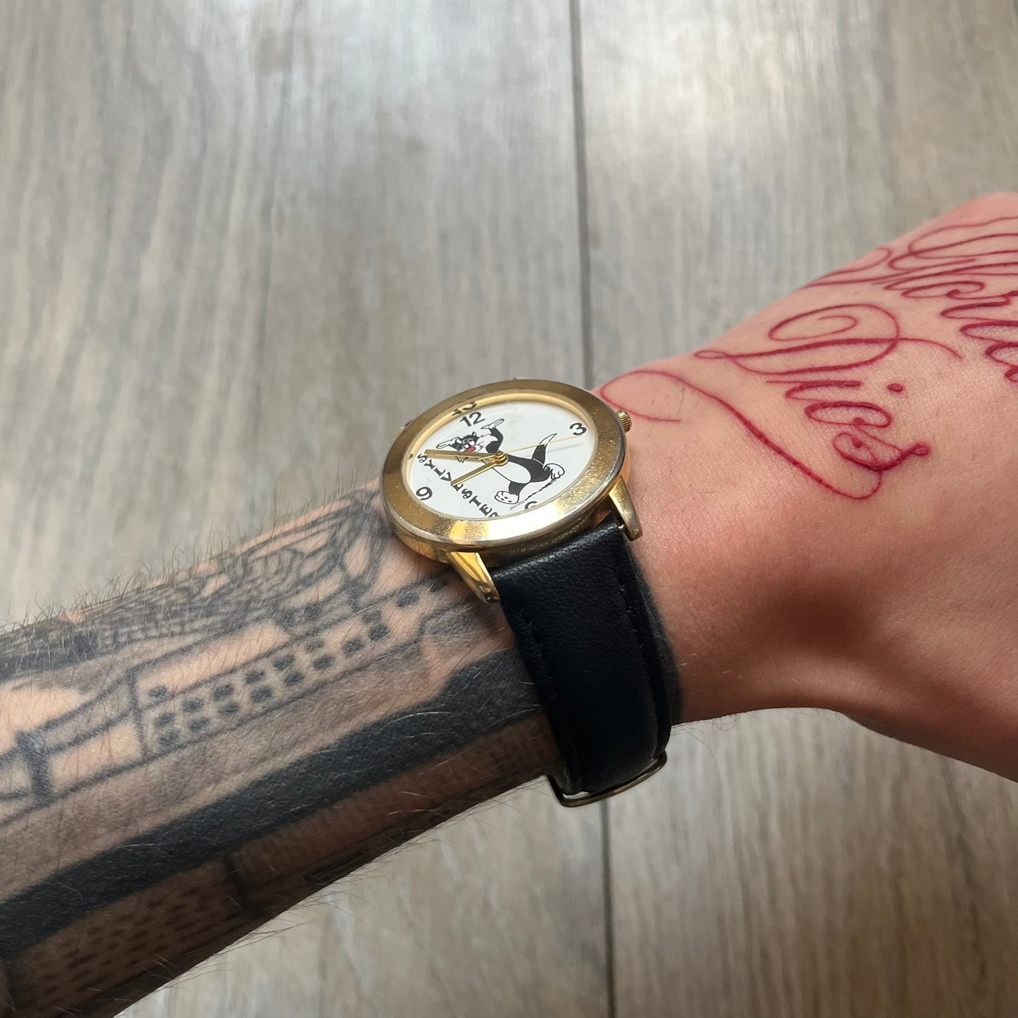 Warner Bros Sylvester 1990 Analog Watch features a gold-tone case with a white dial and black leather strap marked as genuine leather. The dial displays Sylvester with vertical “Sylvester” text, along with hour markers at 12, 3, 6, and 9. The watch uses a Japan movement and includes Warner Bros 1990 licensing engraved on the caseback