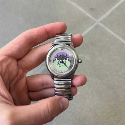 Warner Bros Tasmanian Devil 1996 Analog Watch is a stainless steel watch with a stretch expansion bracelet made from connected metal links instead of a clasp. The watch features a reflective metallic dial with pink triangles and a graphic of Taz smiling in front of a purple background. The caseback includes Warner Bros 1996 licensing.&nbsp;