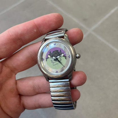 Warner Bros Tasmanian Devil 1996 Analog Watch is a stainless steel watch with a stretch expansion bracelet made from connected metal links instead of a clasp. The watch features a reflective metallic dial with pink triangles and a graphic of Taz smiling in front of a purple background. The caseback includes Warner Bros 1996 licensing.&nbsp;