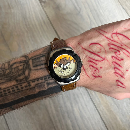 Warner Bros Tasmanian Devil 1997 Analog Watch is a stainless steel watch with a black-silver case and a fresh brown leather strap. The dial features a graphic of Taz smiling in front of a orange background. The caseback includes Warner Bros 1997 licensing