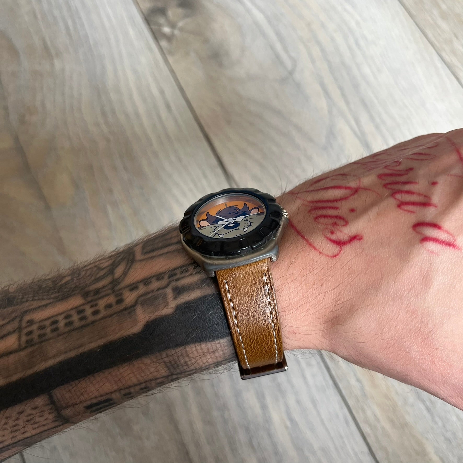 Warner Bros Tasmanian Devil 1997 Analog Watch is a stainless steel watch with a black-silver case and a fresh brown leather strap. The dial features a graphic of Taz smiling in front of a orange background. The caseback includes Warner Bros 1997 licensing