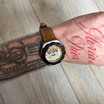 Warner Bros Tasmanian Devil 1997 Analog Watch is a stainless steel watch with a black-silver case and a fresh brown leather strap. The dial features a graphic of Taz smiling in front of a orange background. The caseback includes Warner Bros 1997 licensing