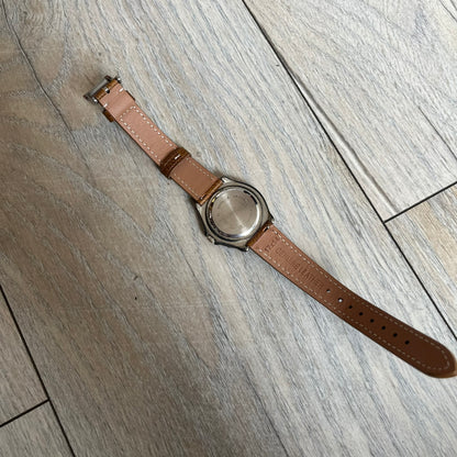 Warner Bros Tasmanian Devil 1997 Analog Watch is a stainless steel watch with a black-silver case and a fresh brown leather strap. The dial features a graphic of Taz smiling in front of a orange background. The caseback includes Warner Bros 1997 licensing