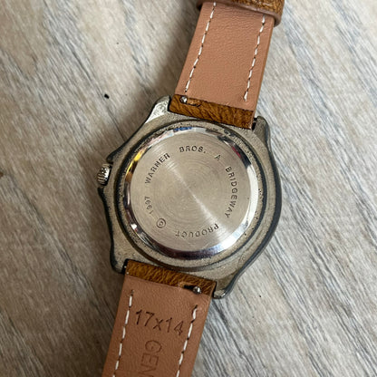Warner Bros Tasmanian Devil 1997 Analog Watch is a stainless steel watch with a black-silver case and a fresh brown leather strap. The dial features a graphic of Taz smiling in front of a orange background. The caseback includes Warner Bros 1997 licensing