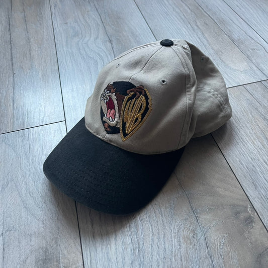 Warner Bros Taz 1998 Baseball Cap is a two-tone beige and black cap featuring an embroidered Warner Bros logo on the front, positioned next to a detailed Taz (Tasmanian Devil) character graphic. Inside, the cap retains its original 1998 Warner Bros tag. The adjustable back strap has been reattached and stitched.