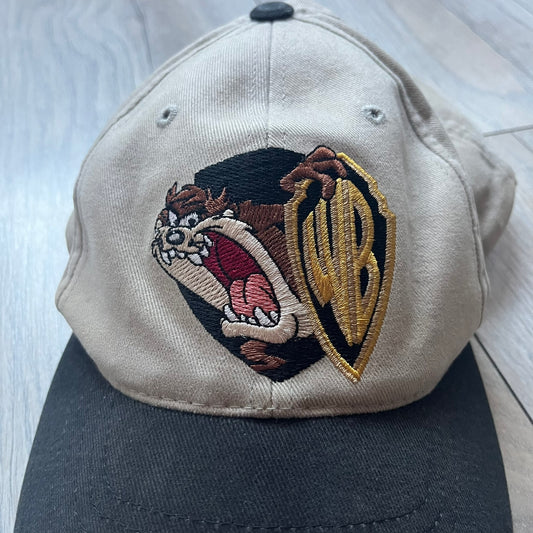 Warner Bros Taz 1998 Baseball Cap is a two-tone beige and black cap featuring an embroidered Warner Bros logo on the front, positioned next to a detailed Taz (Tasmanian Devil) character graphic. Inside, the cap retains its original 1998 Warner Bros tag. The adjustable back strap has been reattached and stitched.