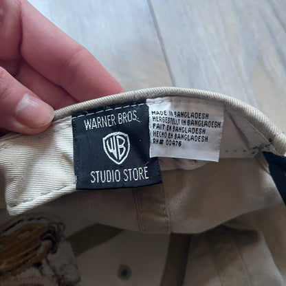 Warner Bros Taz 1998 Baseball Cap is a two-tone beige and black cap featuring an embroidered Warner Bros logo on the front, positioned next to a detailed Taz (Tasmanian Devil) character graphic. Inside, the cap retains its original 1998 Warner Bros tag. The adjustable back strap has been reattached and stitched.