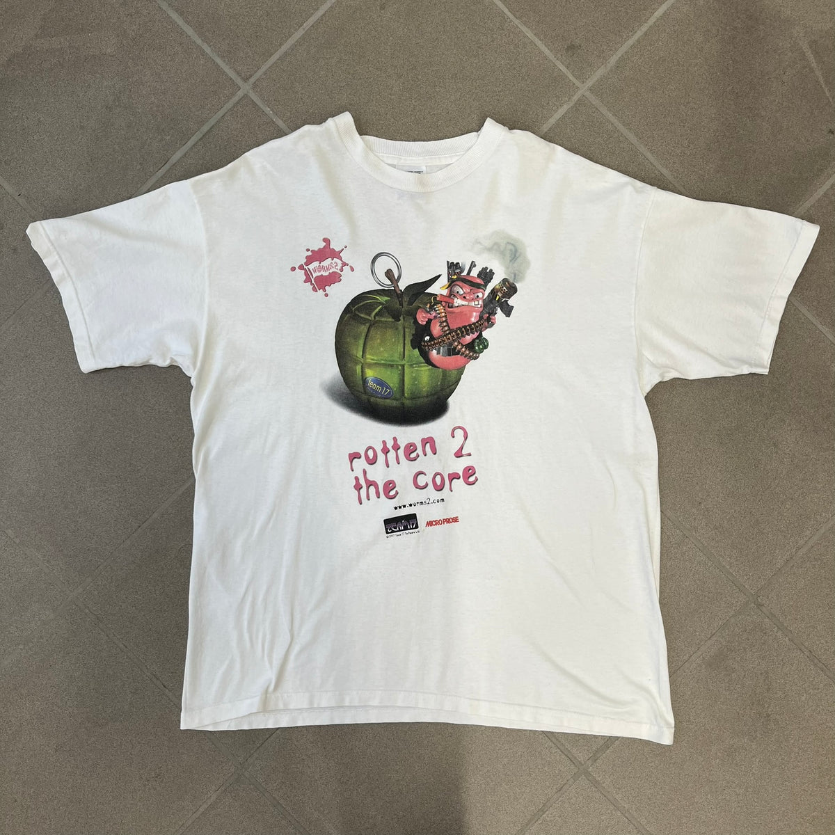 Worms 2 “Rotten 2 The Core” 1997 Tee is a white T-shirt with a front graphic of an apple styled like a grenade, with an angry worm holding a gun coming out of it. The Worms 2 logo sits to the left, and red “Rotten 2 The Core” text below the apple is followed by the website and company logos and a small 1997 licensing mark.