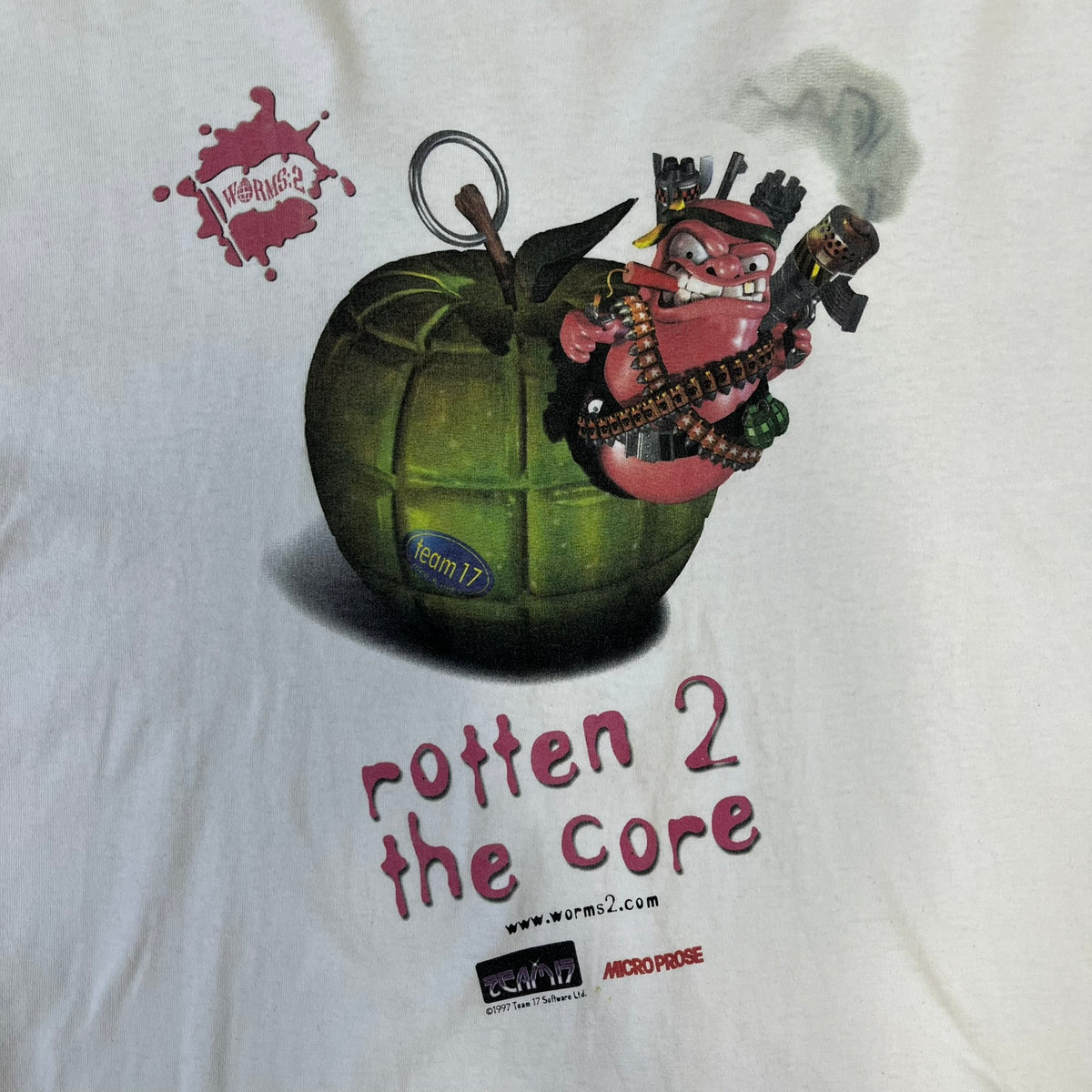 Worms 2 “Rotten 2 The Core” 1997 Tee is a white T-shirt with a front graphic of an apple styled like a grenade, with an angry worm holding a gun coming out of it. The Worms 2 logo sits to the left, and red “Rotten 2 The Core” text below the apple is followed by the website and company logos and a small 1997 licensing mark.