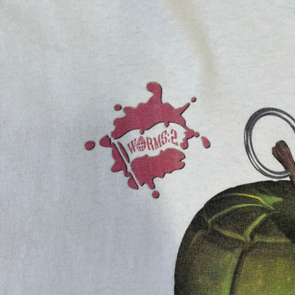 Worms 2 “Rotten 2 The Core” 1997 Tee is a white T-shirt with a front graphic of an apple styled like a grenade, with an angry worm holding a gun coming out of it. The Worms 2 logo sits to the left, and red “Rotten 2 The Core” text below the apple is followed by the website and company logos and a small 1997 licensing mark.